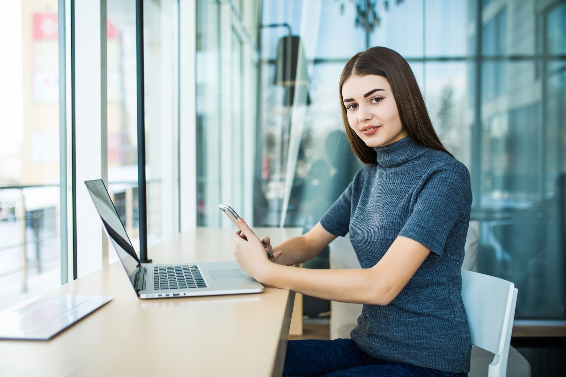 Side view of young smiling businesswoman sitting at table in coffee shop and uses smartphone on table cup of coffee and notebook.